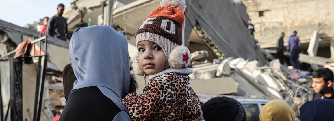 Kleines Kind auf dem Arm vor zerstörten Häusern in Gaza